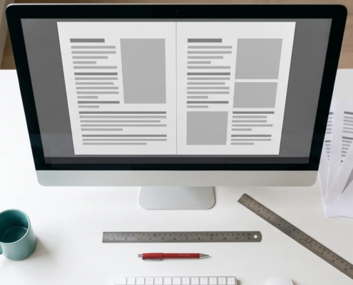 Designer's desk with a monitor showing a right-aligned two-page layout, printed proofs, a ruler and a red proofing pen