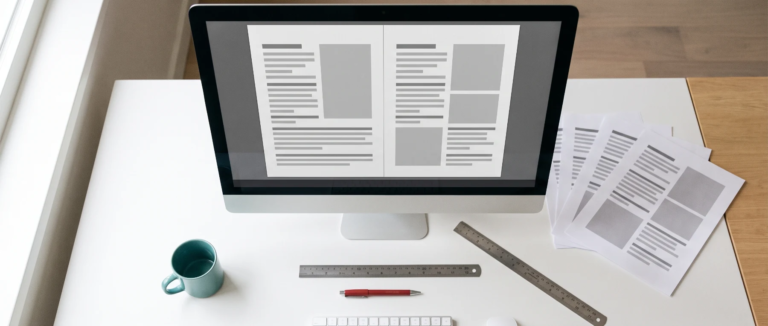 Designer's desk with a monitor showing a right-aligned two-page layout, printed proofs, a ruler and a red proofing pen